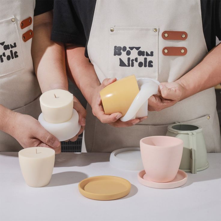 Dos personas vestidas con delantales sostienen moldes de silicona para velas en forma de cuenco, mostrando el proceso de creación de velas minimalistas. En la mesa se observan diferentes piezas, incluyendo velas y tapas en tonos pastel.